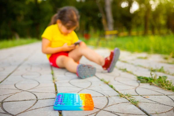 Missbrukare flicka sitter på parken vägen och spela smartphone spel framför sin nya trendiga pop det leksak. — Stockfoto