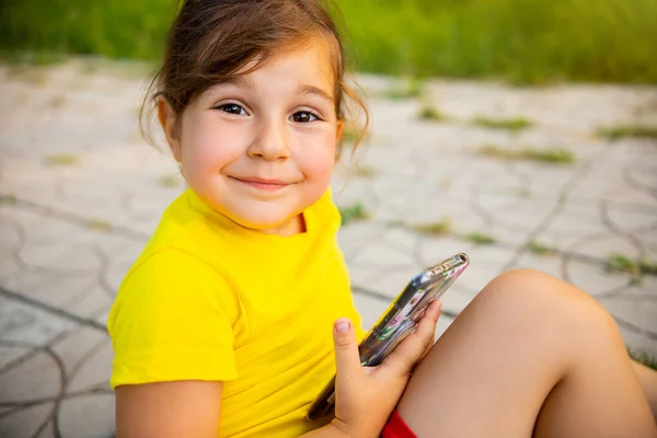 Close up photo of a lovely little girl preschool age being in the park sitting on the path holding the smartphone in her hands and looking straight at the camera. — стоковое фото