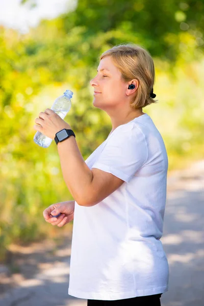 La photo de profil de la femme âgée sportive fatiguée après des exercices de remise en forme a ouvert la bouteille d'eau tenant dans ses mains et va la boire, portant un casque et un téléphone intelligent. — Photo