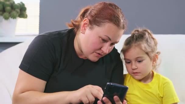 Plus size woman showing to her little daughter information on the smartphone and suddenly mother covers her daughter eyes, so that she does not see bad thinks. — Stock Video