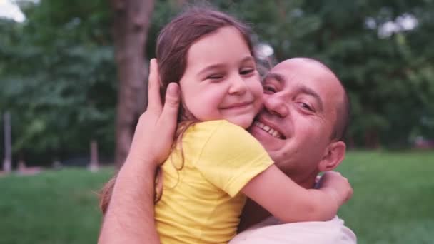 W dzień taty ojciec i jego mała córeczka spacerują po parku, on trzyma ją w rękach całując ją i przytulają się z uśmiechem. — Wideo stockowe