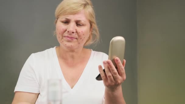 La femme blonde assise à la table se regarde dans le miroir de la main et utilise des bourgeons de coton pour mettre des médicaments contre l'acné sur le visage. — Video