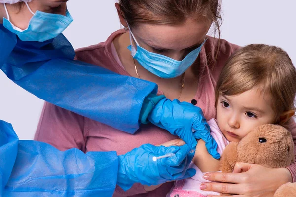 Foto de cerca de una madre en una mascarilla que sostiene a su pequeña niña de rodillas y la médica está poniendo una vacuna a su hija usando jeringa desechable con aguja. — Foto de Stock