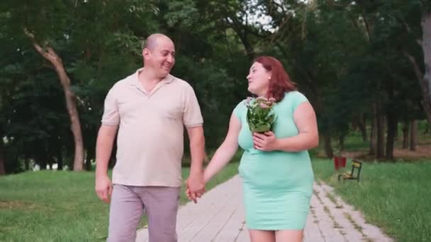 Casal jovem de homem e mulher grávida caminhando explorações mãos no caminho no parque, ela mantém em seu braço o vaso de flores e eles estão falando com um sorriso. — Vídeo de Stock