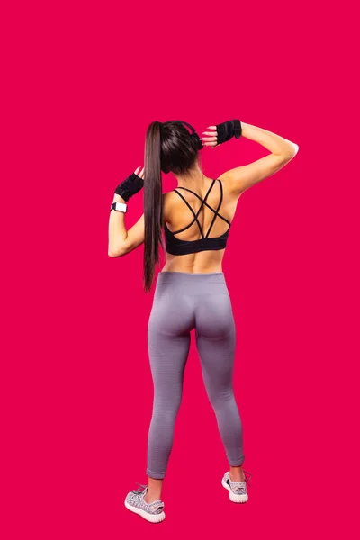Back view of athletic beautiful young sexy woman wearing leggings and short top with cross straps on the back, listening the music using headphones. — Stock Photo, Image