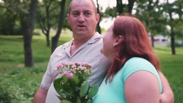 公園を歩いている美しい大人のカップルの男と女性、赤い髪の女性は彼女の腕の中で花を維持し、笑顔. — ストック動画