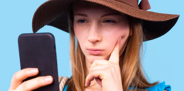 Закрыть фотографию задумчивой блондинки с косоглазыми глазами в коричневой шляпе, смотрящей на телефон, держащий в руке. — стоковое фото