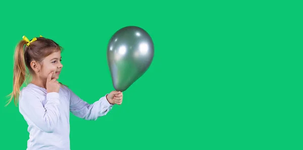 Adorável menina brilhante segurando um balão verde escuro na frente dela e pensando para quem dá-lo. — Fotografia de Stock