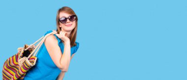 Güneş gözlüklü açık mavi bluz giyen mutlu bir kadın arkasında renkli çizgiler olan çantayı tutuyor..