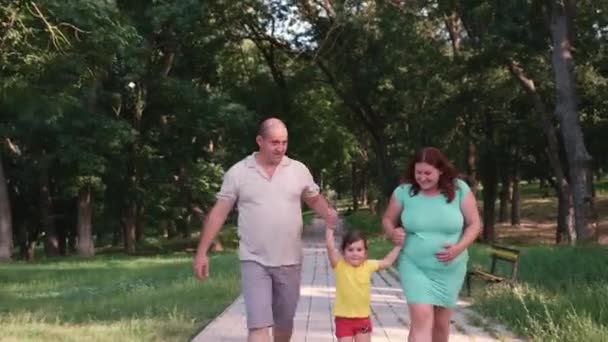 Zwangere vrouw met haar familie wandelen in het park langs het pad houdt het kleine meisje aan de handen en het opvoeden van haar, zwangere vrouw ondersteunt haar buik met haar hand. — Stockvideo