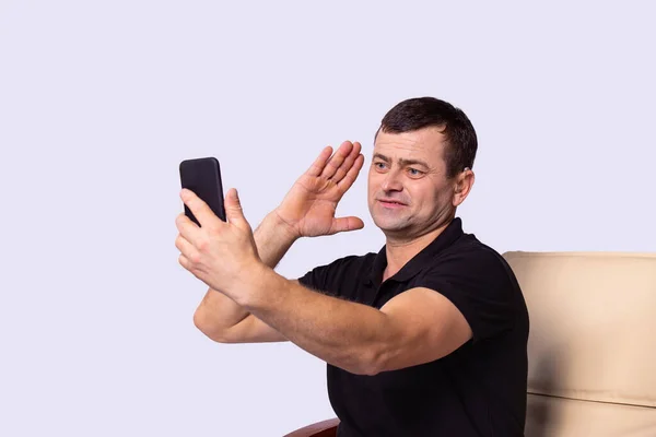 Der Mann im schwarzen T-Shirt sitzt mit Hörgerät auf einem Stuhl, telefoniert per Videotelefon und winkt. — Stockfoto