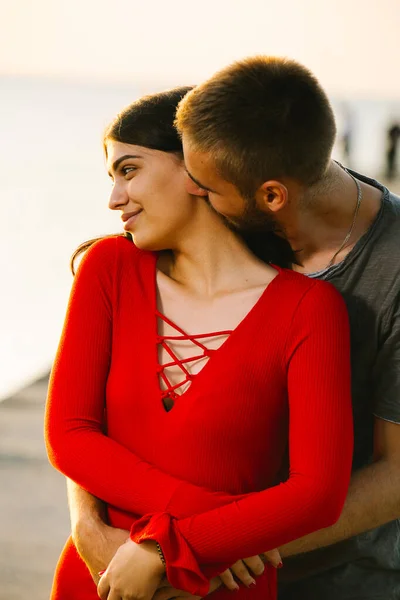 Heureux jeune couple amoureux ayant soirée romantique, le jeune homme étreint sa belle jeune femme en robe rouge par derrière et l'embrasse sur le cou. — Photo