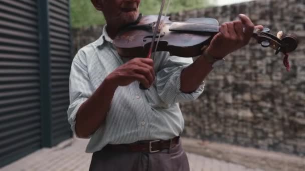 Den närbild video av en gammal man bär vit randig skjorta med långa ärmar och bruna byxor på ett bälte med klockan oh handen spelar fiol mycket professionellt utomhus. — Stockvideo