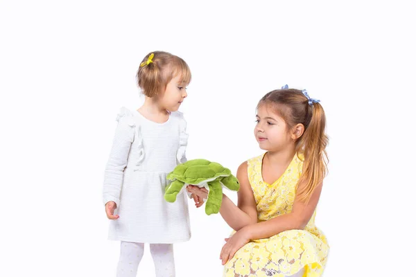 Två söta blonda systrar poserar i vackra klänningar och två hästsvansar, äldre syster håller i handen en mjuk sköldpadda leksak och vill ge den till sin yngre syster. — Stockfoto