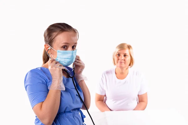 Den kvinnliga läkaren i uniform, medicinsk mask och skyddshandskar gropar på ett stetoskop för att lyssna på patienten andas. — Stockfoto