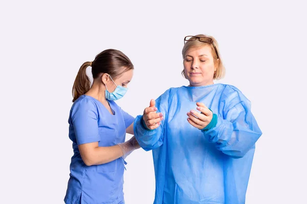 Sjuksköterskan i skyddsmask sätter en skyddande klänning på en blond kvinna med glasögon på huvudet för operation. — Stockfoto