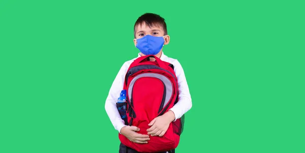 Un enfant en uniforme scolaire portant un masque médical se prépare pour l'école tenant un sac à dos rouge. — Photo