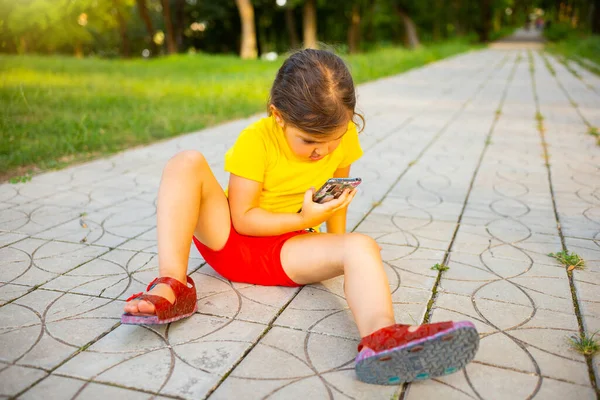 Niña sentada en el camino en el parque vistiendo camiseta amarilla y pantalones cortos rojos manteniendo el teléfono inteligente en sus manos y mirándolo. —  Fotos de Stock
