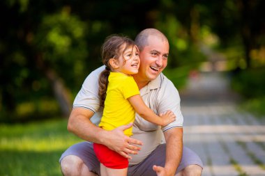 Küçük kızıyla dizlerinin üzerine çömelmiş mutlu baba gülümsüyor ve onu sevgiyle kucaklamak istiyor. Parkta birlikte iyi vakit geçirmek istiyor..