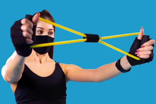 Close up of a fitness young girl using sports elastic rubber trying to stretch it wearing textile black mask in pandemic time. — Stock Photo, Image