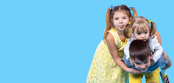 Tre unga söta barn som har roligt visar tungan flicka i gul klänning och en pojke som håller på sin rygg lilla flicka — Stockfoto