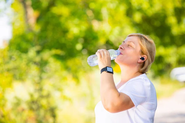 Belle femme blonde sportive portant des écouteurs et montre intelligente numérique a une pause après avoir fait du jogging à l'extérieur dans le parc, l'eau potable de la bouteille en plastique. — Photo