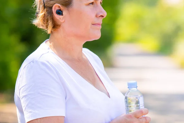 La photo de profil d'une femme adulte blonde sportive portant un appareil auditif et tenant la bouteille d'eau dans ses mains, se reposant après avoir fait du jogging à l'extérieur. — Photo