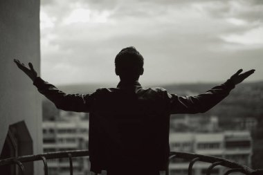 Siyah beyaz bir fotoğraf ceketli bir adamın balkonda dikilirken kollarını açmış özgürlüğünü hissederken..