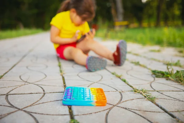 Suddig bild av en liten flicka i gul t-shirt och röda shorts sitter på vägen i parken och hålla den smarta telefonen i händerna och spela, pop det leksak är nere på vägen, beroendeframkallande koncept. — Stockfoto