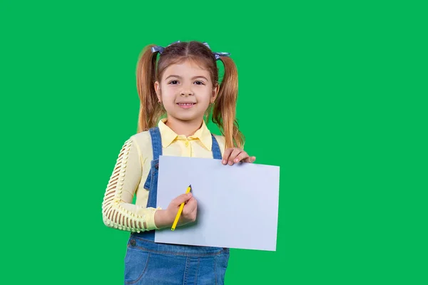 Έξυπνο κοριτσάκι που κοιτάζει την κάμερα κρατώντας ένα λευκό χαρτί και ένα στυλό στα χέρια της και αρχίζει να γράφει. — Φωτογραφία Αρχείου
