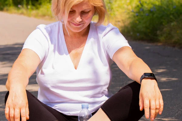 Heureuse femme blonde âgée confiante méditant en position lotus sur la route dans le parc portant une montre intelligente sur sa main et une bouteille d'eau . — Photo