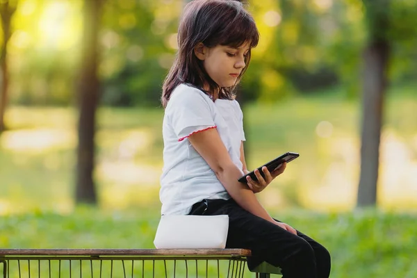 Vue de côté photo d'une jolie jeune fille assise sur un banc tenant le téléphone dans sa main et naviguant des informations intéressantes. — Photo