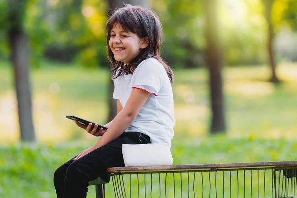 Vista lateral de cerca foto de una chica alegre sentada en un banco con las piernas colgando sosteniendo el teléfono inteligente en una mano y ella se ríe con los ojos cerrados. —  Fotos de Stock