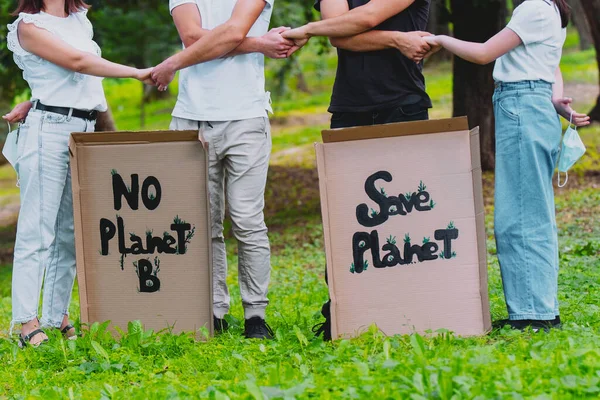 Cropped photo of two couples outdoors holding hands with cardboards posters with the inscription about saving nature, two young women keep medical masks in the hands behind their backs.