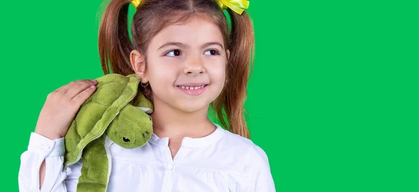 Banner, långt format. Glädjande skolåldern flicka med två hästsvansar bär vit t-shirt håller en mjuk grön sköldpadda leksak på axeln och tittar bort. — Stockfoto