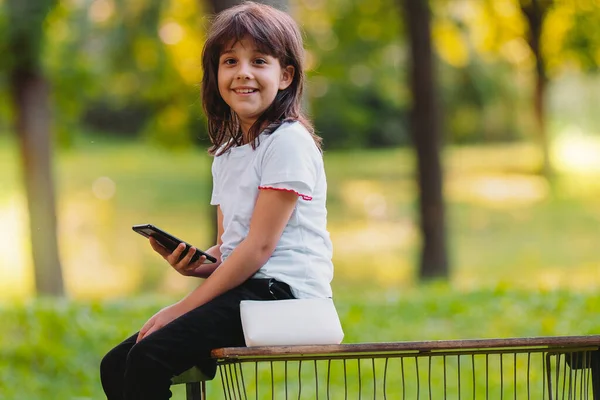 Vue latérale photo d'une fille heureuse assise sur le bord d'un banc regardant la caméra, tenant le téléphone intelligent et sac à main blanc dans ses mains. — Photo