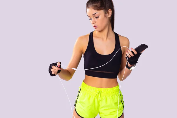 Vonzó fitness fiatal nő áll rövid top és sport rövidnadrág kiegyenesíti a vezetéket a fejhallgató és tartja a telefont a bal kezében. — Stock Fotó