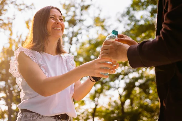Joyeux jeune femme bénévole en chemisier blanc regardant le sans-abri et lui donnant une bouteille d'eau en plastique à boire. — Photo