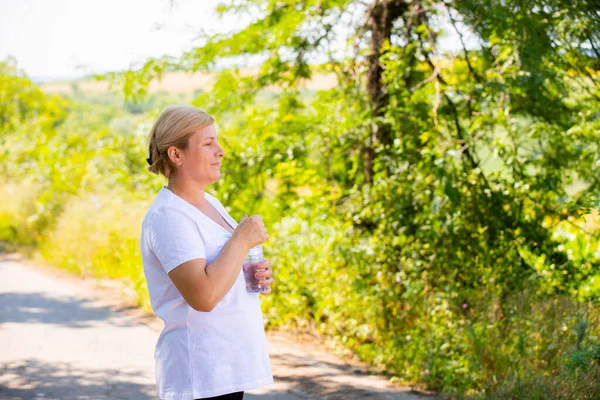 Photo de profil d'une femme sportive âgée debout et se reposant après le jogging matinal dans le parc, tente d'ouvrir une bouteille d'eau qu'elle tient dans ses mains. — Photo