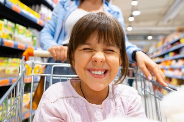 Gülümseyen geniş bir kızın, dişlerini market arabasında annesiyle alışveriş yaparken gösteren yakın bir fotoğrafı. Yüksek kalite fotoğraf