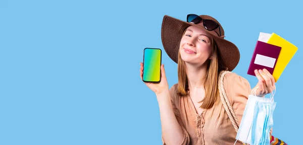 Mujer muy contenta turista con sombrero y gafas de sol posando en cámara con teléfono móvil, máscara médica y pasaporte. — Foto de Stock