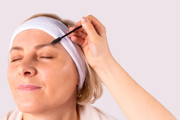Mulher loira em um salão de beleza com cabeça branca passa por um procedimento para pentear as sobrancelhas por um esteticista usando uma escova de sobrancelha. — Fotografia de Stock