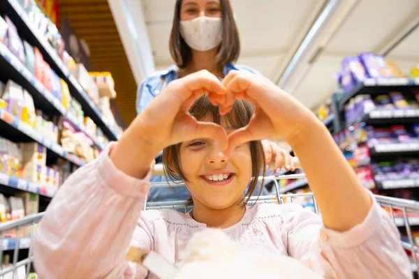 Το όμορφο κορίτσι που κάθεται στο καλάθι με τα ψώνια κάνει σημάδι καρδιάς με τα χέρια της να είναι στην αγορά του καταστήματος, η μητέρα της στέκεται πίσω της με μια ιατρική μάσκα. — Φωτογραφία Αρχείου