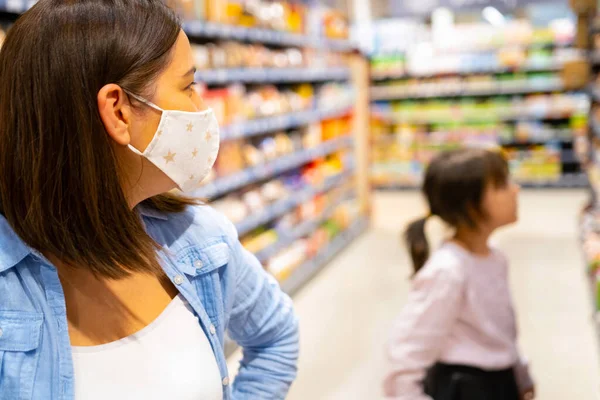 Vista lateral de la foto de dos mujeres mamá persona en una máscara médica y la hija dentro de la tienda mirando los estantes con productos. — Foto de Stock