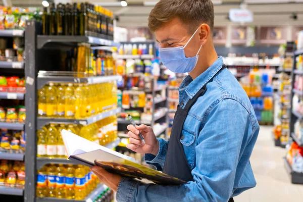 Πλάγια άποψη φωτογραφία ενός ευτυχισμένου εργάτη καταστήματος φορώντας ιατρική μάσκα, denim πουκάμισο και μαύρη ποδιά κρατώντας το ημερολόγιο και κάνοντας απογραφή. — Φωτογραφία Αρχείου