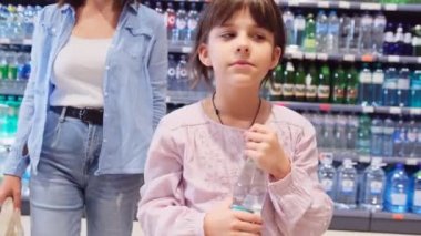 Elinde su şişesini tutan kız annesiyle alışverişe çıkıyor. Elinde tutacağı tutacağı olan çanta, arka planda şişeler olan raflar..