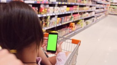 Arkadaki fotoğrafta, alışveriş arabasında oturan esmer bir kız var. Elinde yeşil ekran, elinde akıllı telefon ve rafların arasında yürüyen biri var..