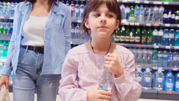 Девушка держит бутылку воды в руках ходить по магазинам со своей матерью за держа сумку с ручками для продуктов, полки с бутылками фона. — стоковое видео