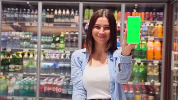 Zárjuk le a videót egy csinos fiatal hölgyről a boltban szódával a háttérben, kezében az okostelefonnal, akinek zöld képernyős hívásai vannak, miközben gesztikulál a nagy hüvelykujjával.. — Stock videók