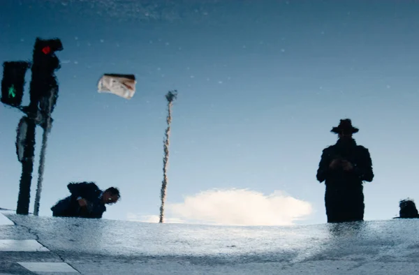 Mavi gökyüzünün üzerinde küçük bir bulutla yansıtıldığı su havuzu, iki iyi giyimli adam, biri çömelmiş, diğeri ise bir şapka ve kırmızı ve yeşil trafik ışığı ile duruyor..